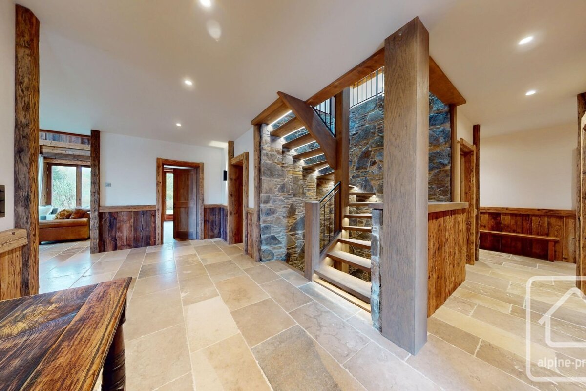 A rustic modern hallway with a lighted wooden staircase against a stone wall, complemented by wooden beams, stone tile floors, and white walls.