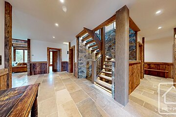 A rustic-modern interior featuring stone floor tiles, wooden beams and paneling, and an illuminated stone and wood staircase. Another room with a sofa is visible.