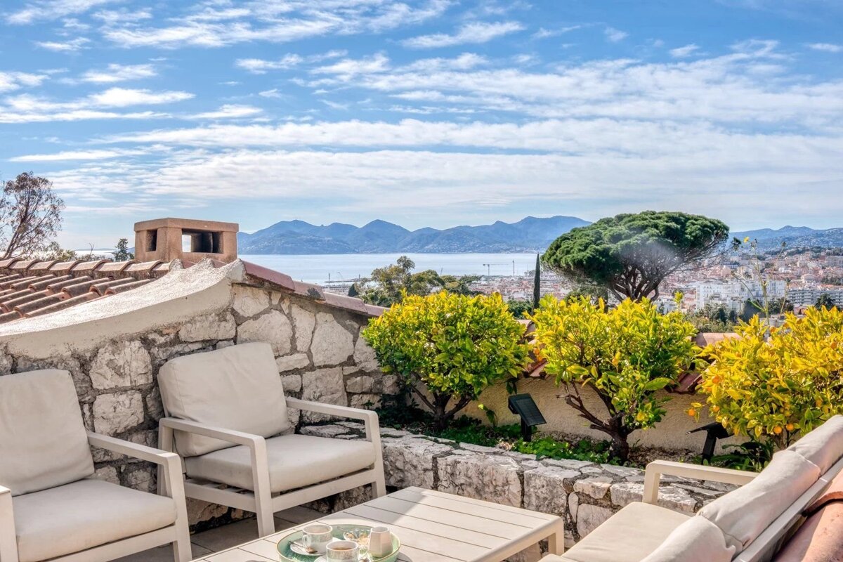 A cozy Mediterranean terrace with armchairs overlooking a coastal city, sea, and distant mountains.
