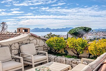 A beautiful Mediterranean patio with outdoor seating, citrus trees, and a stunning view of the sea, mountains, and a coastal town under a blue sky.