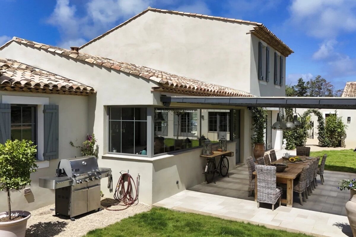 Sunny backyard patio of a stucco house with an outdoor dining table, wicker chairs, a stainless steel grill, and a green lawn.