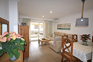 Bright apartment living room with a dining area, beige sofas, wooden furniture, potted plants, and a balcony visible through sliding doors.