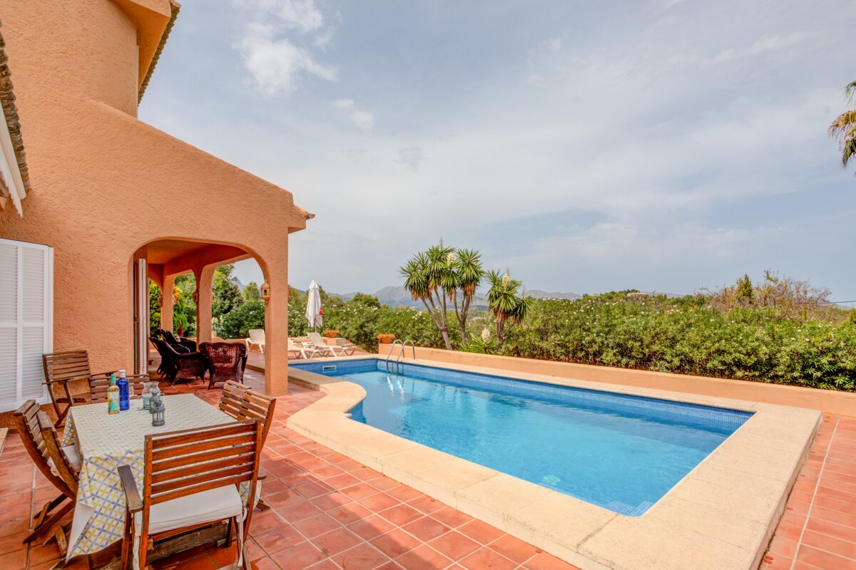 A serene villa patio features a refreshing blue swimming pool, outdoor dining area, and lush green landscaping with scenic distant views.