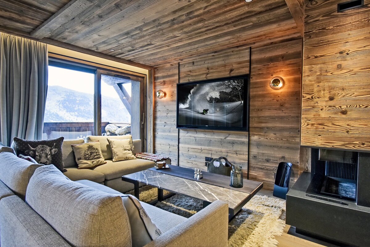 Cozy ski chalet living room with rustic wood paneling, grey sofas, a marble coffee table, and scenic mountain views outside.