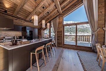 A rustic wooden chalet kitchen with a modern island and stools, offering panoramic snowy mountain and forest views through large windows and a balcony.