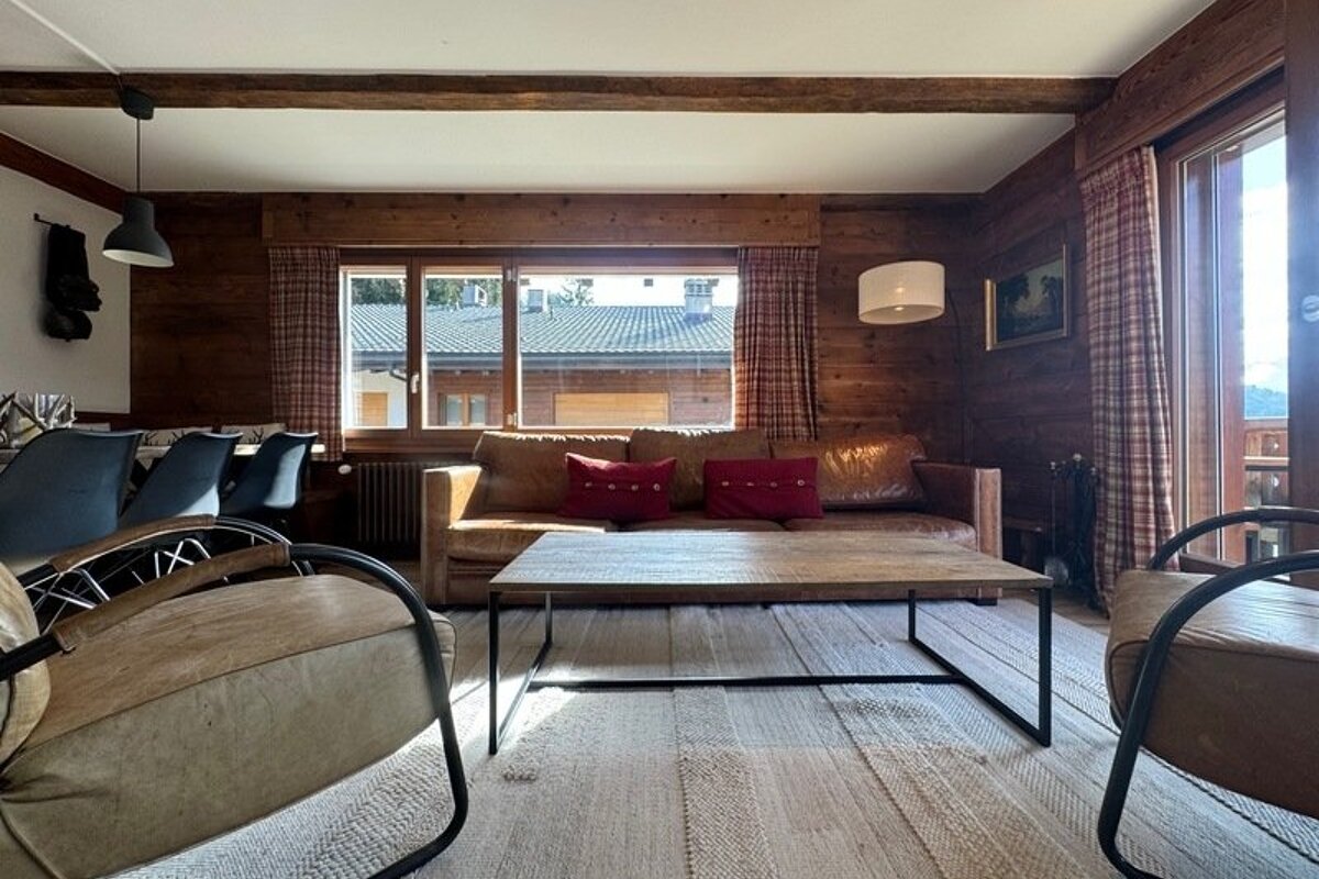 A cozy, rustic living room with wood-paneled walls, a leather sofa with red cushions, a coffee table, and two matching armchairs.