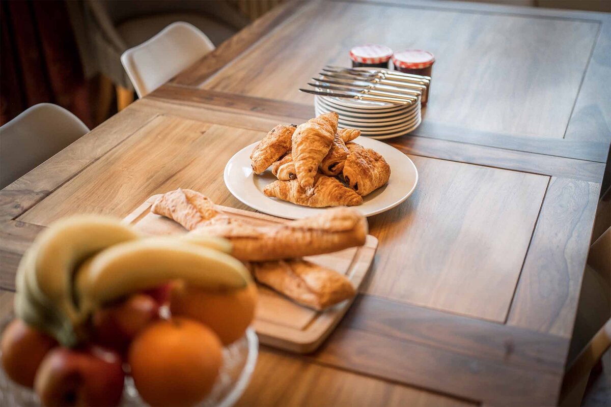 A plate of croissants sits on a wooden table next to a bowl of fruit