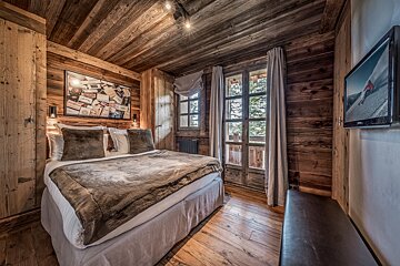 A charming, wood-paneled bedroom with a large bed, a window revealing trees, and a wall-mounted TV showing skiing.