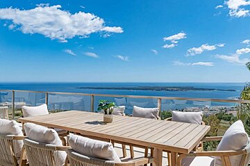 An outdoor dining terrace with a wooden table and chairs, offering panoramic views of a sparkling blue ocean, distant island, and clear sky with white clouds.