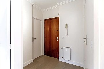 A simple white hallway with grey wood flooring, white doors, a warm wooden front door, and a wall-mounted phone and heater.