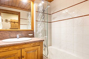 A chalet-style bathroom featuring a wooden vanity with a sink, a mirror, and a shower-bathtub combo behind a decorative glass screen.