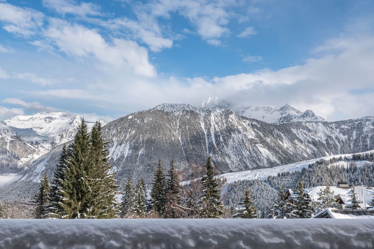 A breathtaking snowy mountain range rises under a partly cloudy blue sky, framed by snow-covered evergreen trees and a thick layer of fresh snow.