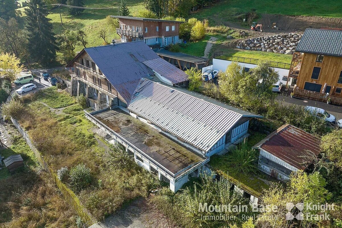 Aerial view of multiple buildings on a grassy hillside with trees and parked cars. Some roofs are weathered, suggesting a rural or mountain property.