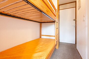 A natural wood bunk bed with an orange mattress on the lower bunk, positioned in a simple room next to a white door and textured wall.