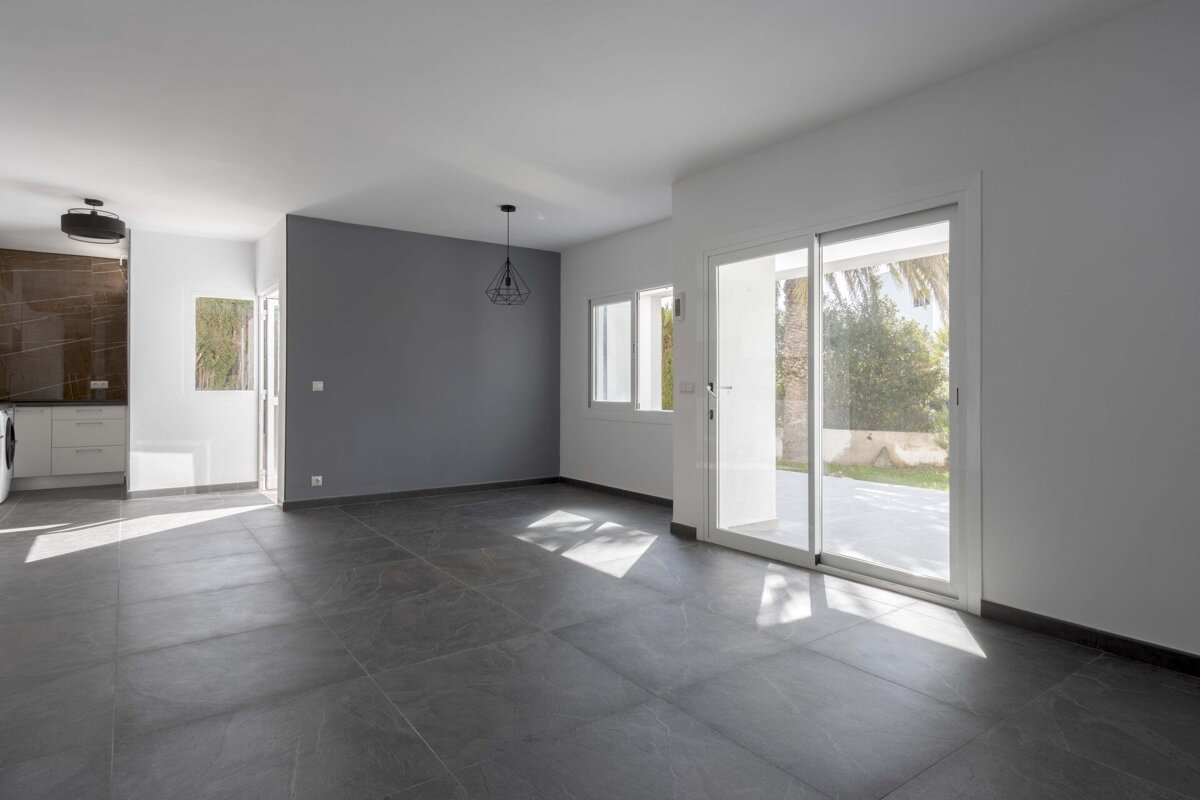 Bright, empty modern room with grey tiled floor, white and grey walls, a kitchenette, and sliding glass doors to a sunny patio.