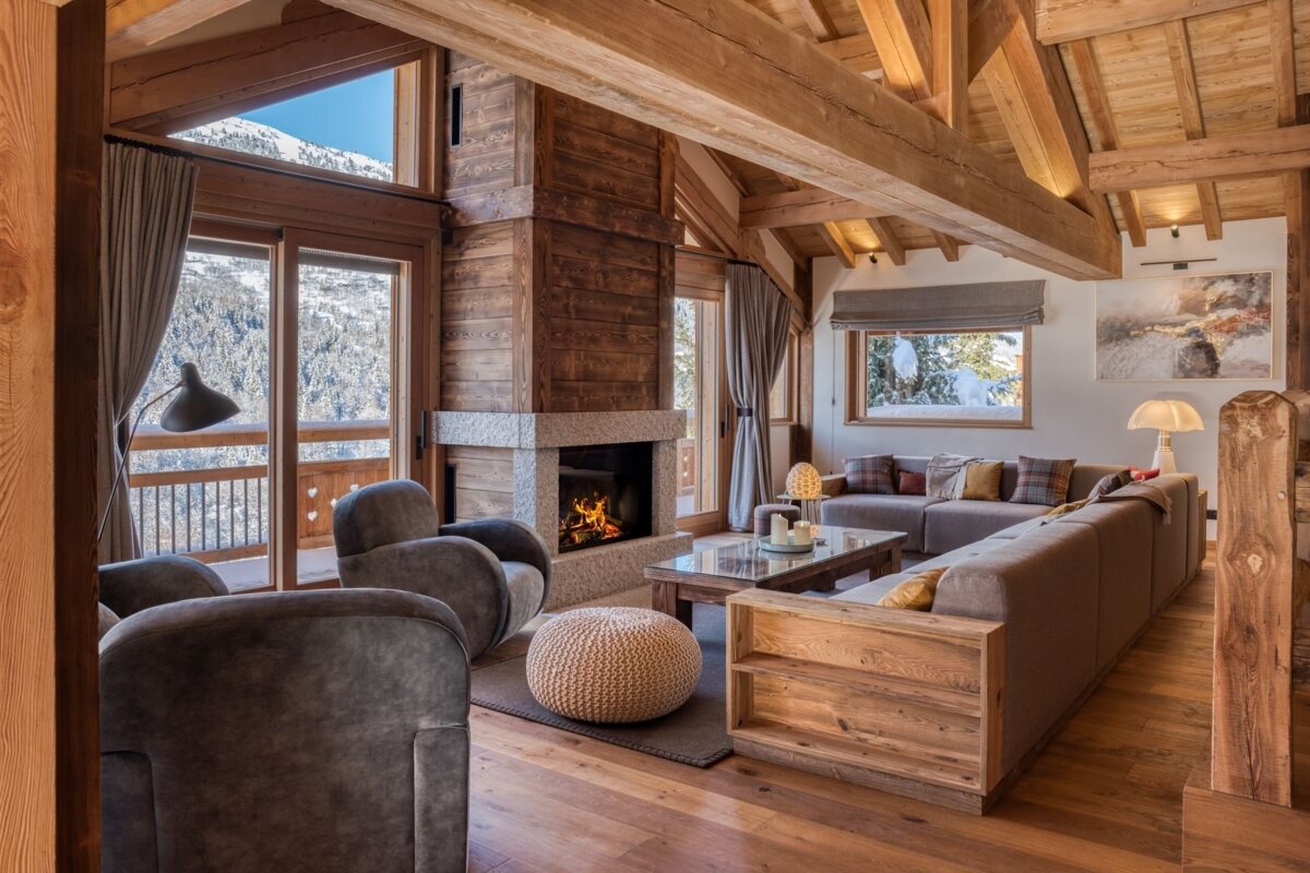 A luxurious ski chalet living room with a cozy fireplace, exposed wooden beams, plush seating, and panoramic snowy mountain views.