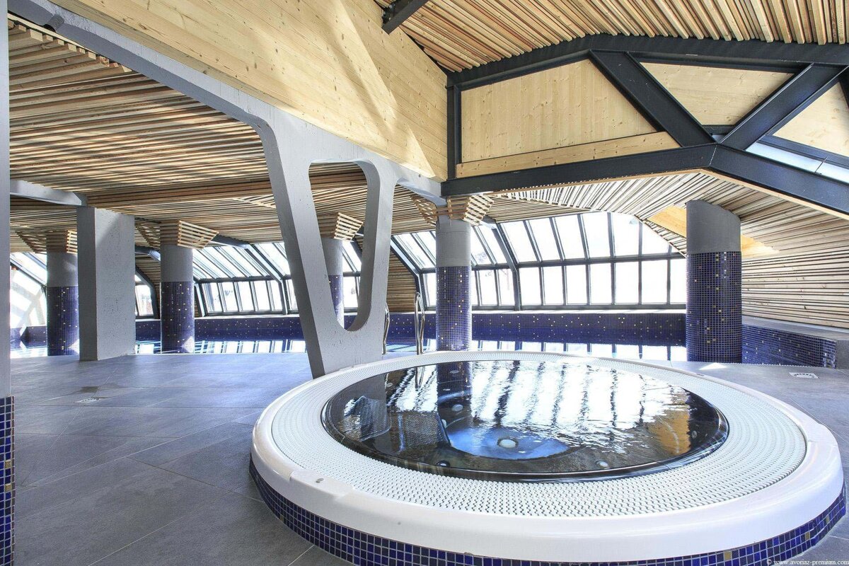 Modern indoor pool and jacuzzi area with wood slat ceiling, large windows, blue mosaic tiles, and striking grey architectural supports.