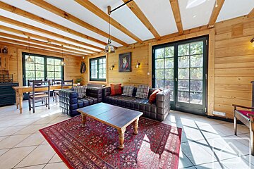 A cozy, rustic room with wooden walls and beamed ceiling. Features brown leather sofas, a red patterned rug, a wooden coffee table, and natural light.