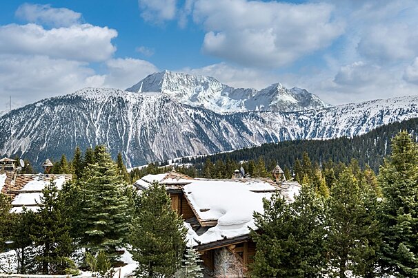 Snow-covered mountains rise above pine forests and rustic chalets, bathed in sunlight under a blue, cloudy sky.