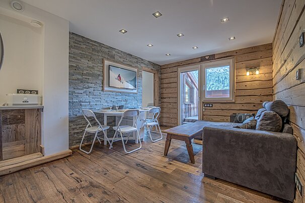 Cozy chalet living/dining space with wood and stone finishes, a dining table, sofa, and balcony overlooking mountains.