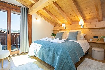 A cozy wooden chalet bedroom with a beamed ceiling, a bed dressed in teal, and glass doors opening to a sunny balcony.