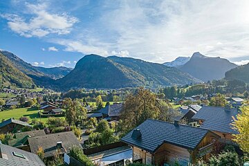 An alpine village nestled in a green valley, surrounded by forested mountains with distant snow-capped peaks under a bright, partly cloudy sky.