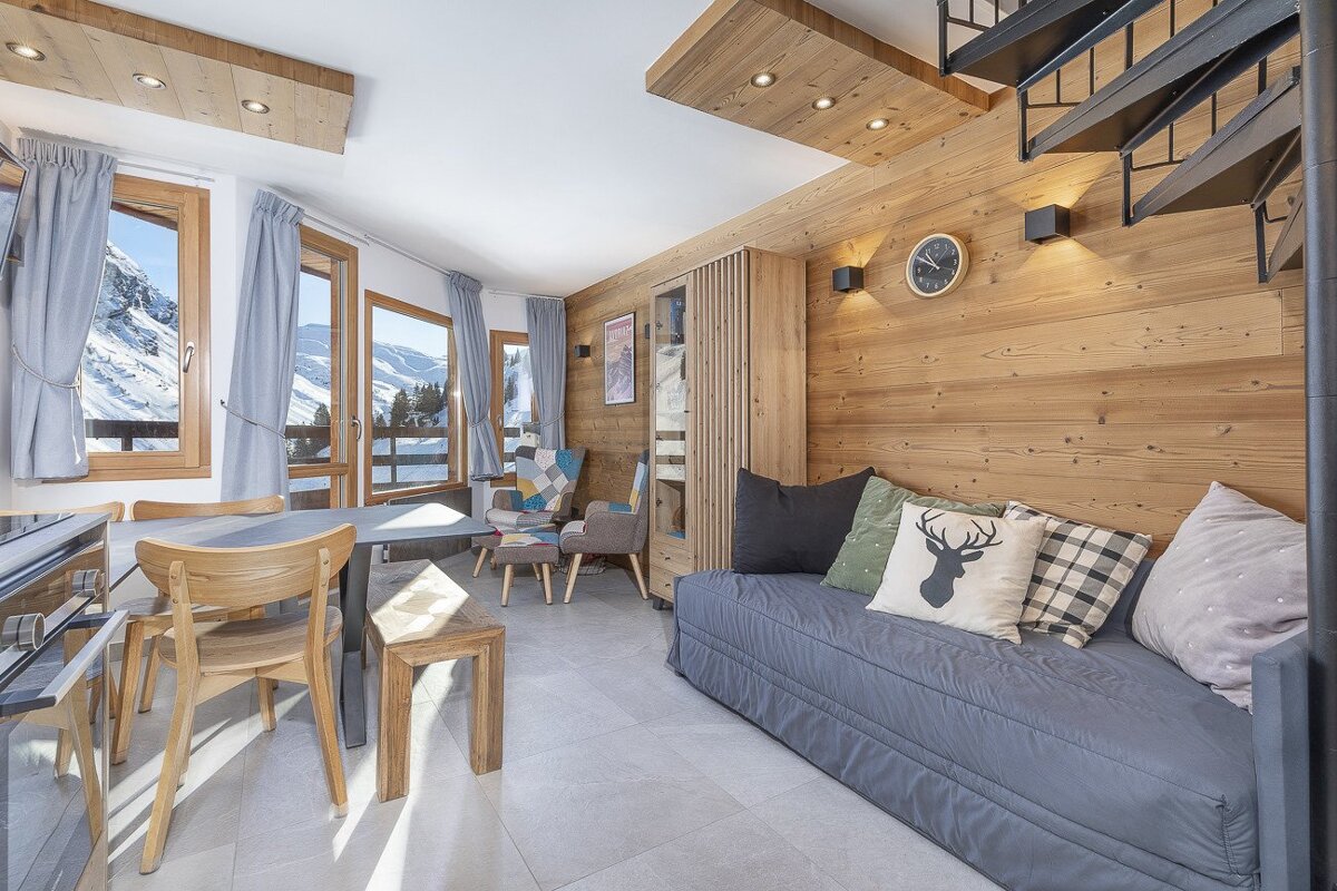 Cozy cabin living room with dining area, sofa, and spiral stairs, boasting large windows with stunning snowy mountain views.