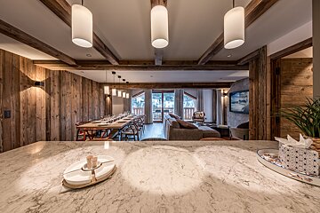 A modern alpine chalet interior with wood-paneled walls, seen from a marble countertop looking towards a dining table and cozy living area.