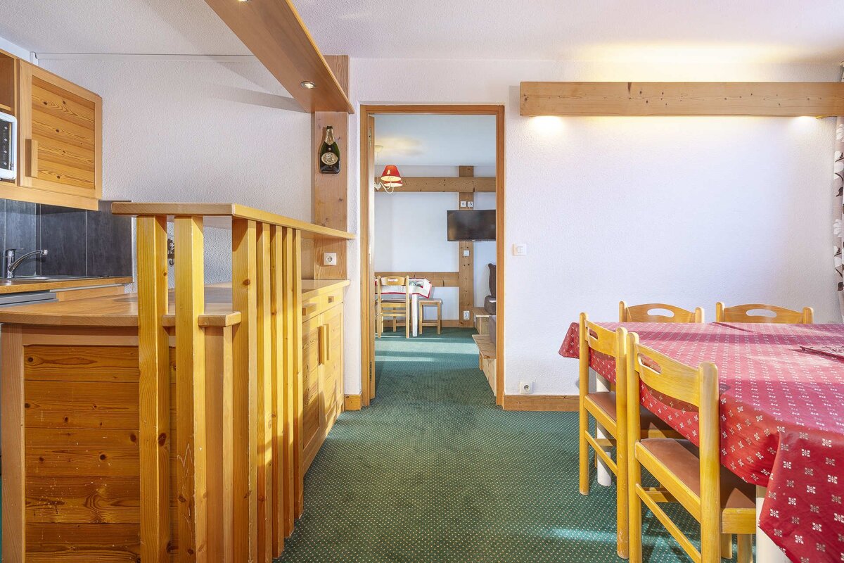 A cozy, wood-paneled interior features a kitchen bar, dining table with a red tablecloth, and green carpet. A doorway leads to another room.