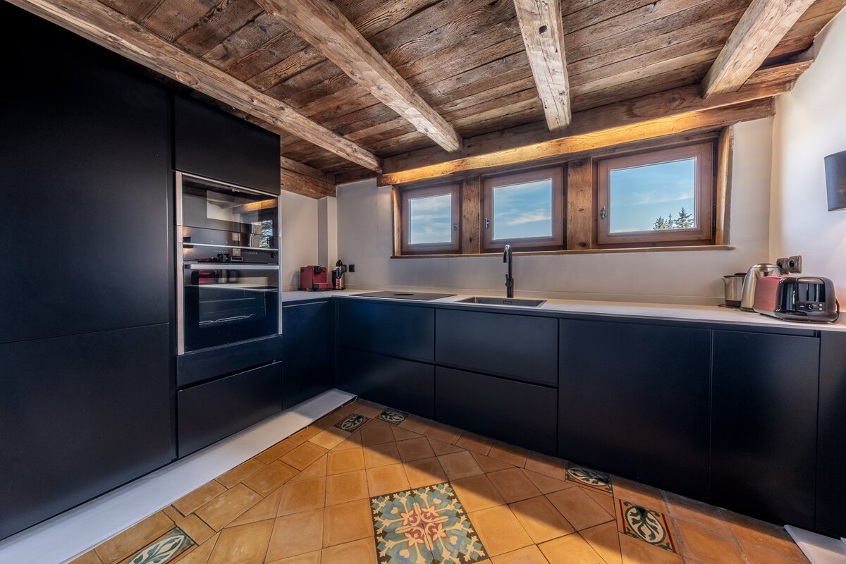 Sleek black kitchen with a rustic wood-beamed ceiling, three small windows, and a decorative tile floor.