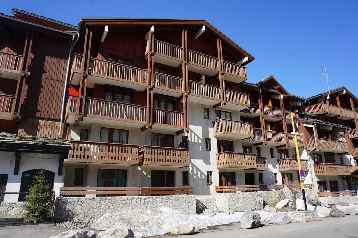A multi-story alpine-style building with wooden balconies and trim, set against a clear blue sky with patches of snow on the ground.