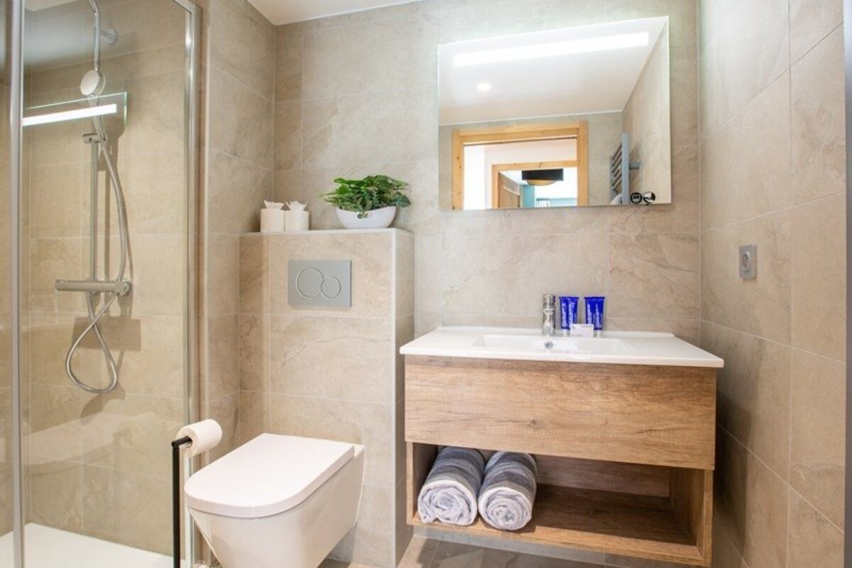 A modern beige-tiled bathroom featuring a glass shower, toilet, and wooden vanity with a sink, mirror, and rolled towels.