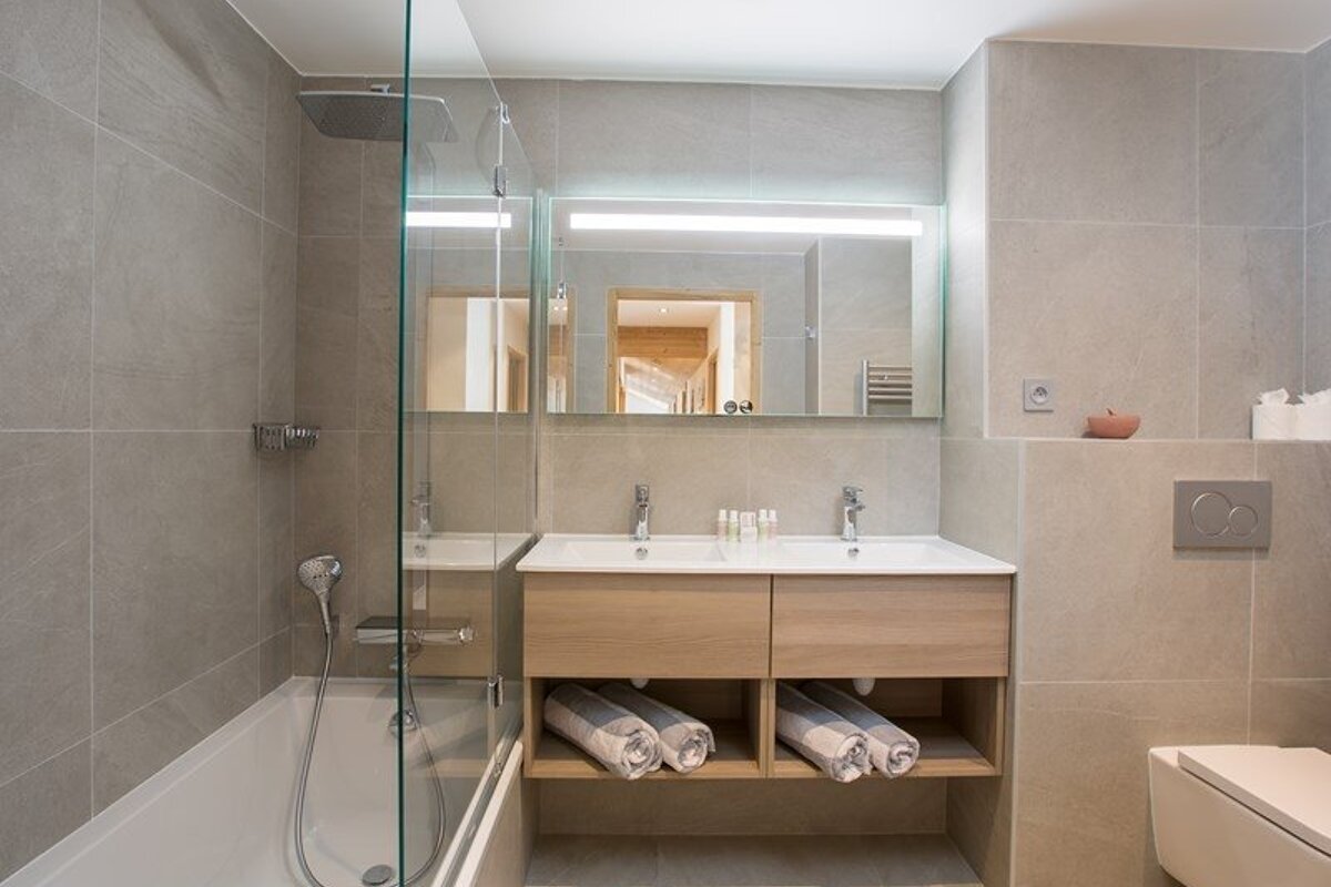 Modern gray-tiled bathroom with a double wooden vanity, a large lit mirror, and a bathtub with a glass screen.