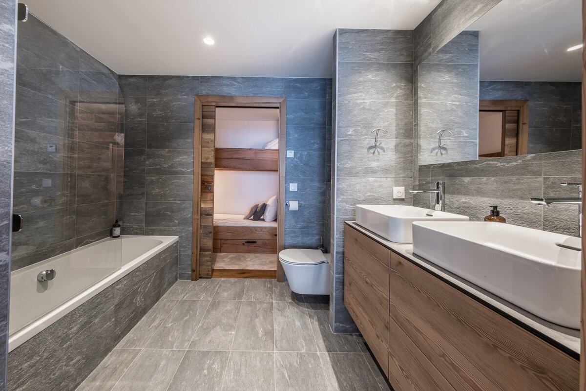 Modern grey-tiled bathroom featuring a double sink vanity, a bathtub, and a doorway leading to a bunk bed room.
