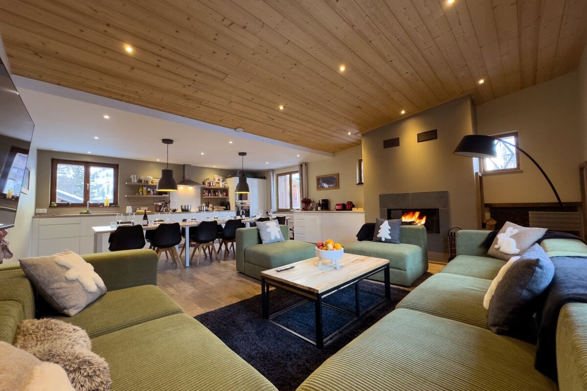 Cozy chalet interior with green corduroy sofas, a lit fireplace, wooden ceiling, and an open kitchen and dining area in the background.