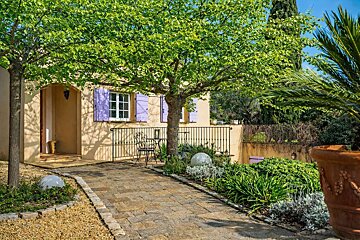 A sun-drenched yellow house with purple shutters, shaded by lush trees, features a stone path leading to a patio with seating in a vibrant garden.
