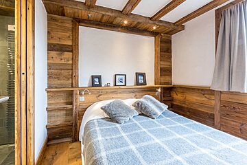 A cozy, rustic bedroom features warm wood paneling, exposed ceiling beams, and a bed with a gray plaid blanket, exuding a chalet-style charm.