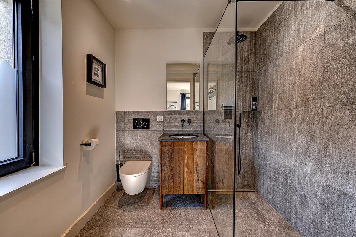 Modern gray-tiled bathroom with a toilet, wooden vanity, sink, and a glass-enclosed walk-in shower.