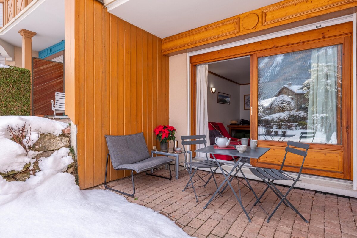 A cozy winter patio with snow on the ground, featuring an outdoor dining set and a sliding door reflecting a snowy mountain view.