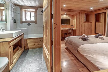 A cozy, wood-paneled chalet-style bedroom with a bed, desk, and TV, openly connected to a bathroom with a tub, sink, and tiled floor.