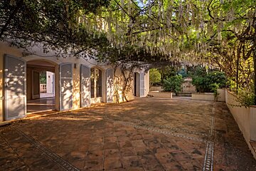 Sunlit tiled patio of a villa, featuring a lush green trellis overhead with hanging white flowers.