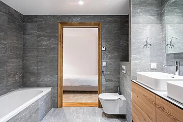 Modern grey-tiled bathroom with a bathtub, toilet, wooden vanity, and doorway leading to a bedroom.