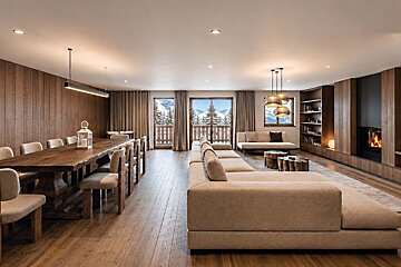 Luxurious chalet interior featuring a large wooden dining table, a plush sectional sofa, a fireplace, and snowy mountain views.