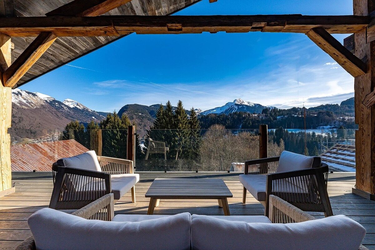 Rustic wooden balcony with modern outdoor furniture frames a stunning view of snowy mountains, pine forests, and a bright blue sky.