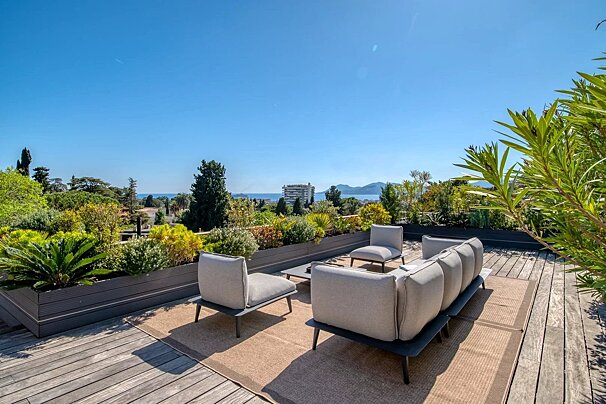 Sunny rooftop terrace with wooden decking, plush outdoor seating, lush green planters, and a scenic sea view under a clear blue sky.