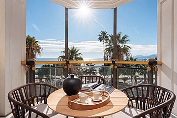 Sunny balcony with coffee, a book, and wicker chairs, offering a beautiful view of the ocean, palm trees, and city beyond.