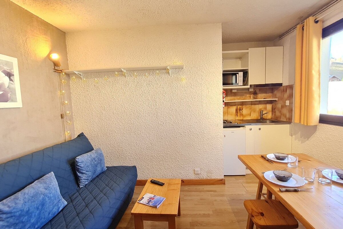 A cozy studio apartment featuring a blue sofa, wooden coffee and dining tables, and a compact kitchenette. Soft lighting and a window view complete the space.