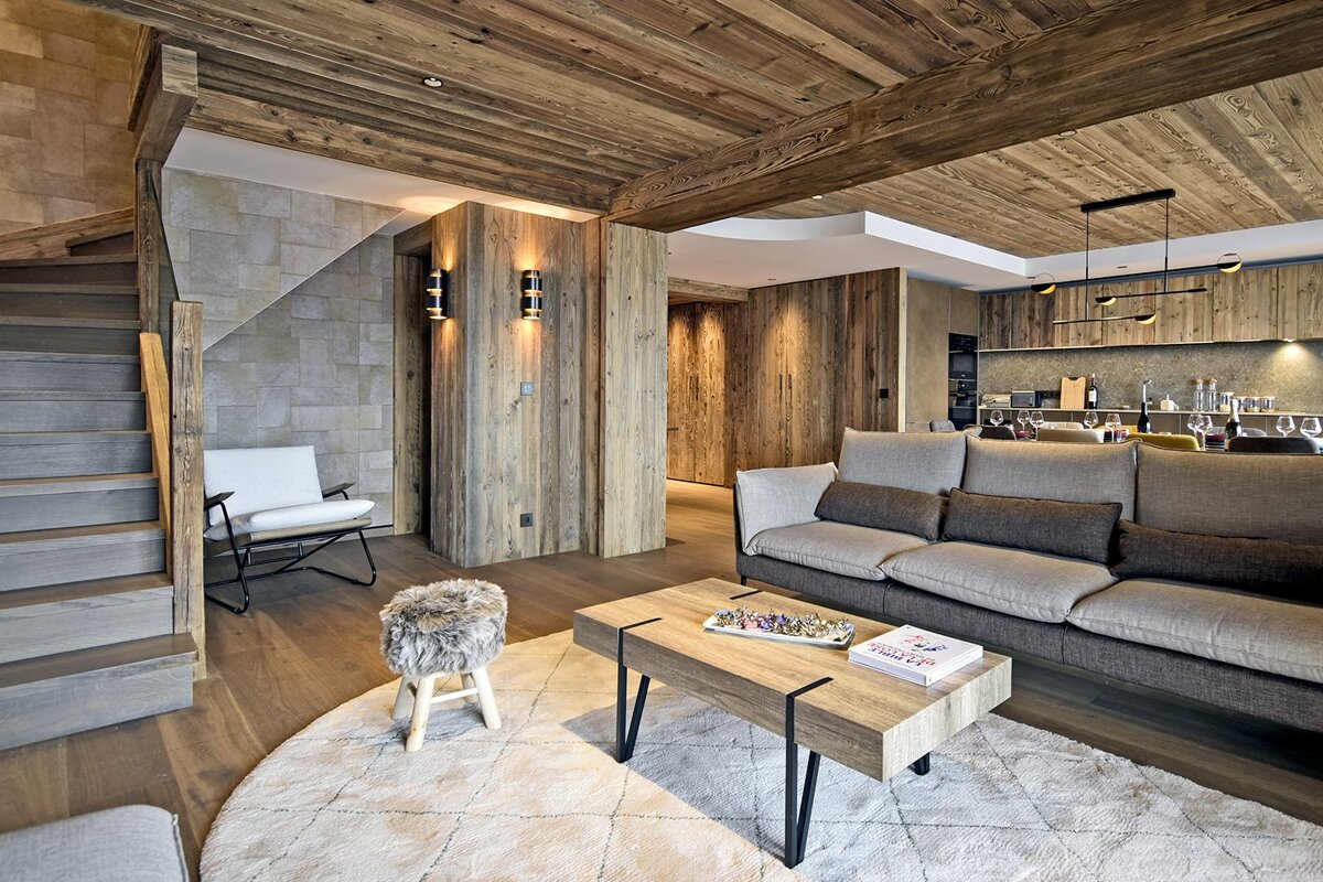 Cozy chalet living room featuring wood-clad walls, a large grey sofa, a wooden coffee table on a white rug, and a staircase.
