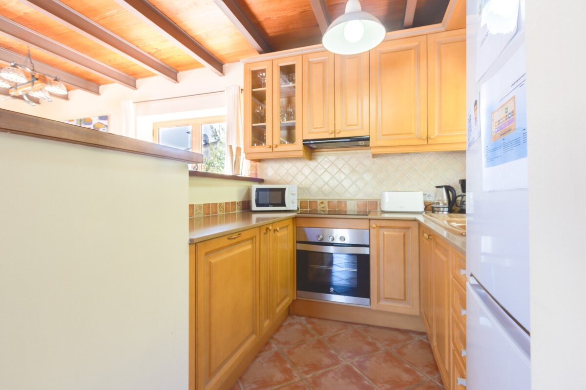 A bright kitchen with light wood cabinets, terracotta floor tiles, exposed wooden ceiling beams, and appliances including a microwave, oven, and fridge.