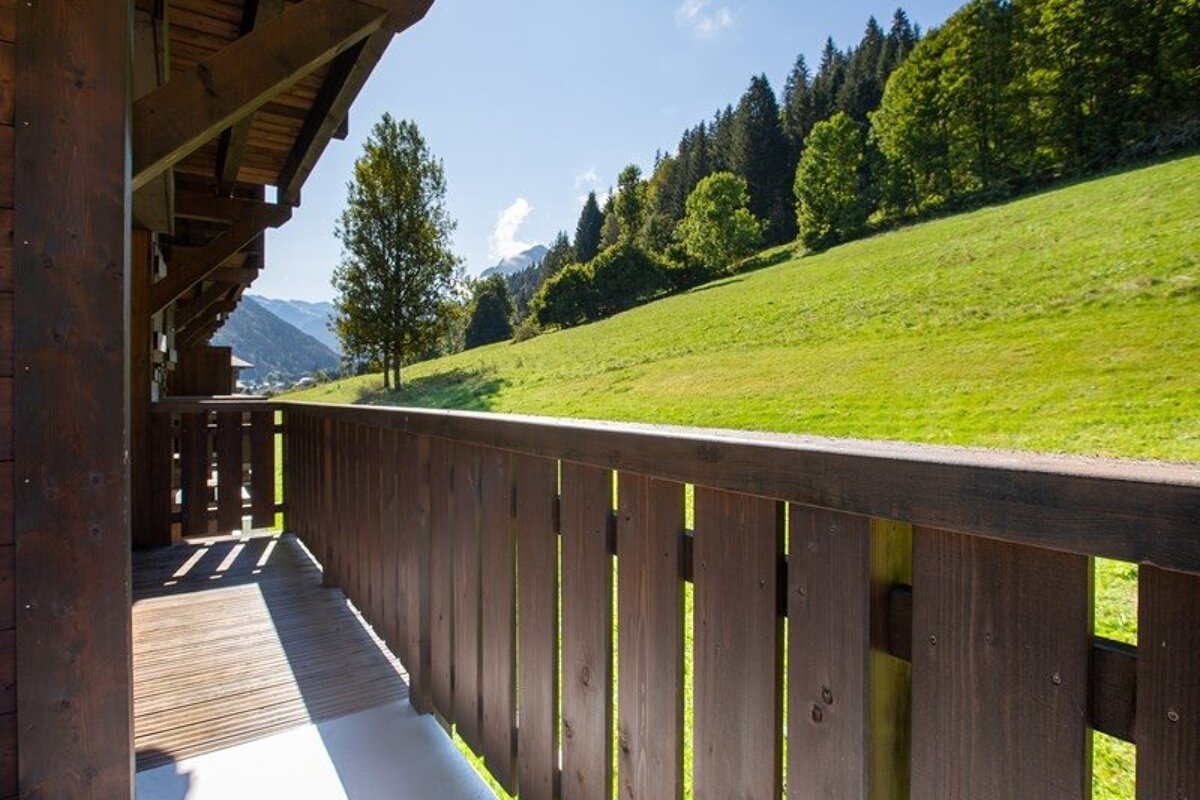 View from a wooden chalet balcony overlooking a lush green hillside and pine forest under a clear blue sky.